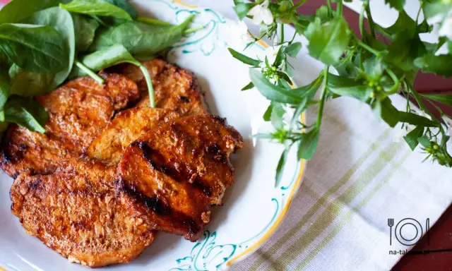 Grillowane plastry schabu w aromatycznej marynacie, podane z zielonymi liśćmi na talerzu.