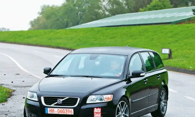 Czarne Volvo V50 2.0D na mokrej drodze, za kierownicą siedzi mężczyzna. W tle zielone wzgórza i budynek.