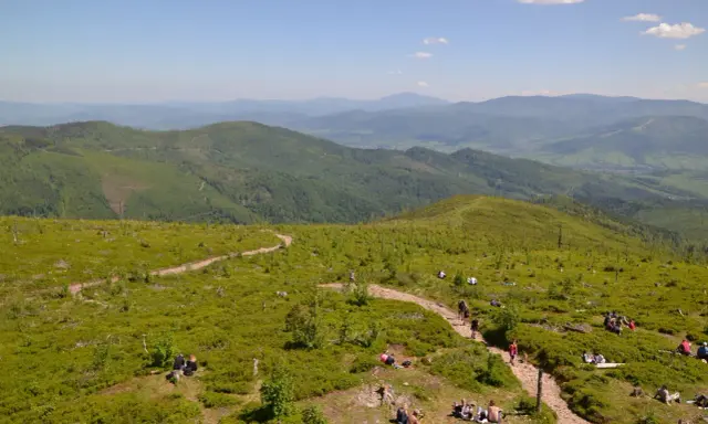 Malownicze widoki na Beskid Śląski, gdzie liczne istebne szlaki turystyczne prowadzą przez zielone wzgórza pełne jagód. Ludzie odpoczywają na trawie, ciesząc się słońcem.