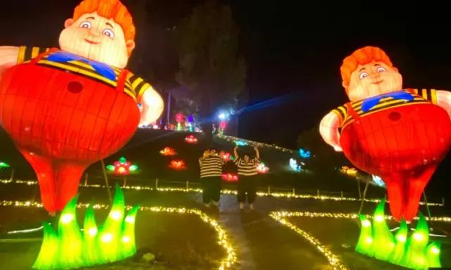 Gigantesche lanterne di bambini paffuti illuminano un sentiero a Wonderland Rimini, con due persone che salutano tra fiori luminosi.