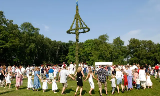 Människor dansar runt en midsommarstång i en grönskande park. Många barn med blomsterkransar i håret deltar i firandet.