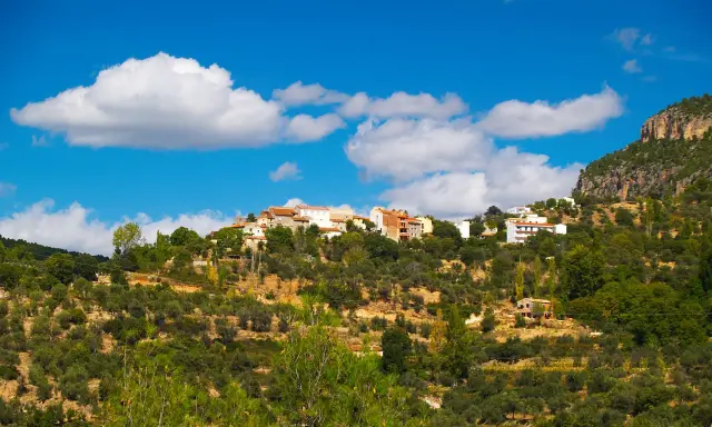 Pueblo blanco en la ladera de una montaña, rodeado de olivos y vegetación. El cielo azul con nubes blancas corona la vista de Gontar, Albacete.