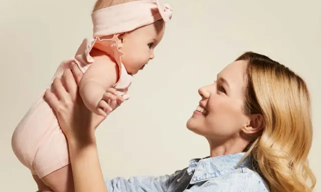 Mama podnosi dziecko w różowym body i opasce. Idealny przykład, jak ubierać noworodka latem – lekko i wygodnie.