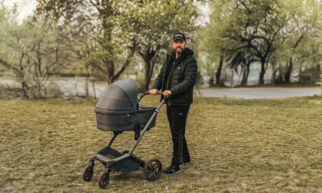 Mężczyzna w czapce z daszkiem i kurtce pcha nowoczesny wózek dla dziecka po trawie.