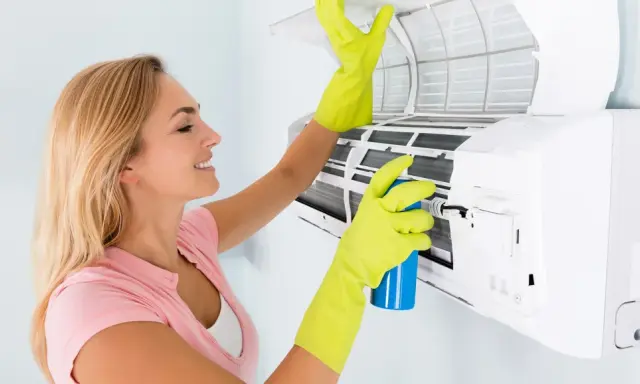 Mujer con guantes verdes limpia el aire acondicionado que huele a humedad, rociando un producto.