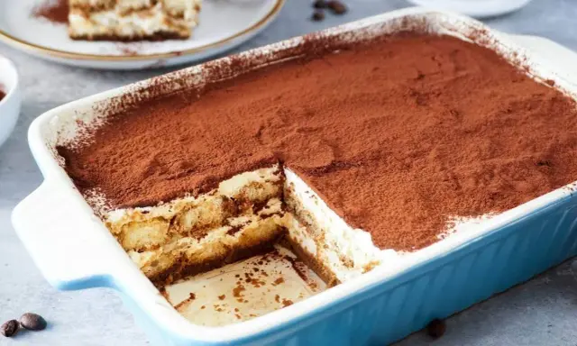 Ein köstliches Tiramisu ohne Ei, in einer blauen Auflaufform, bestäubt mit Kakao. Ein Stück ist bereits entnommen.