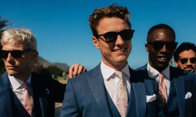 Four men in blue suits with pink ties and sunglasses, embodying a "dress to impress" dress code for a sunny outdoor event.