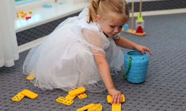 Mała dziewczynka w białej sukience bawi się klockami domino, tworząc kącik zabaw dla dzieci na weselu.