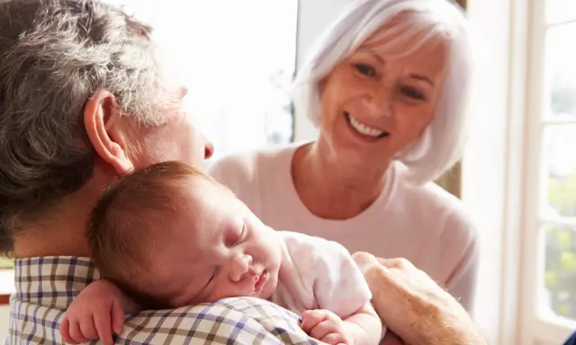 Los abuelos paternos sonríen mientras el abuelo sostiene al bebé dormido en su hombro.