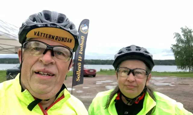 Två cyklister redo för Vätternrundan. Mannen har en keps med texten "Vätternrundan". Bakom dem syns en sjö och en flagga för Sollers IF Fotboll.