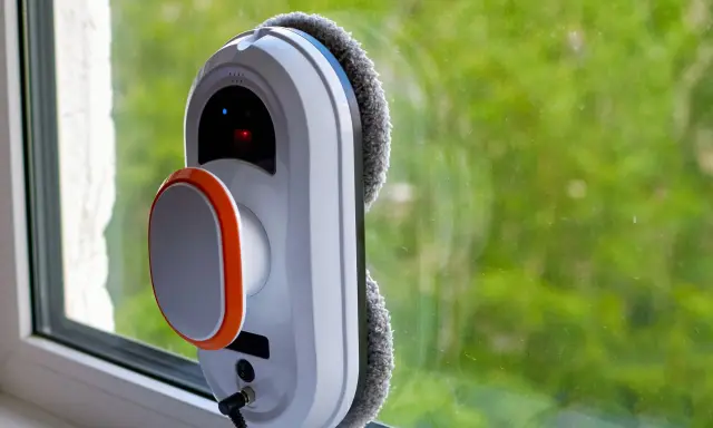 Robot do mycia okien przyczepiony do szyby, z widokiem na zielone drzewa.