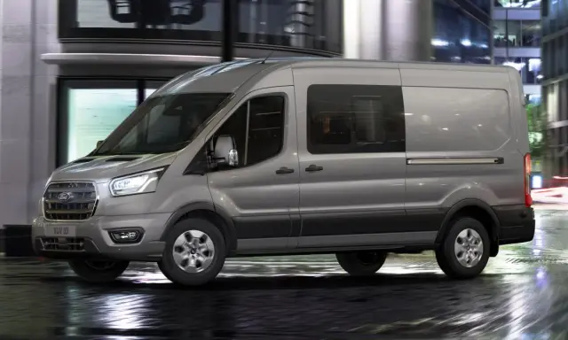 Camión N1 gris Ford Transit circulando por una calle urbana mojada de noche, con luces de edificios y tráfico difuminadas al fondo.