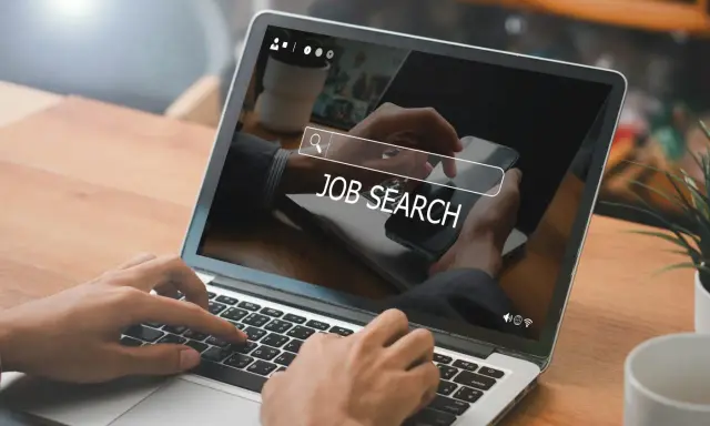 Ręce wpisują "JOB SEARCH" na laptopie. Praca wre, szukamy nowych możliwości.