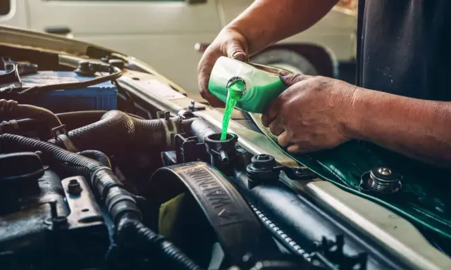 Mecánico rellenando refrigerante verde en el radiador de un coche, solucionando una posible fuga de gas aire acondicionado.