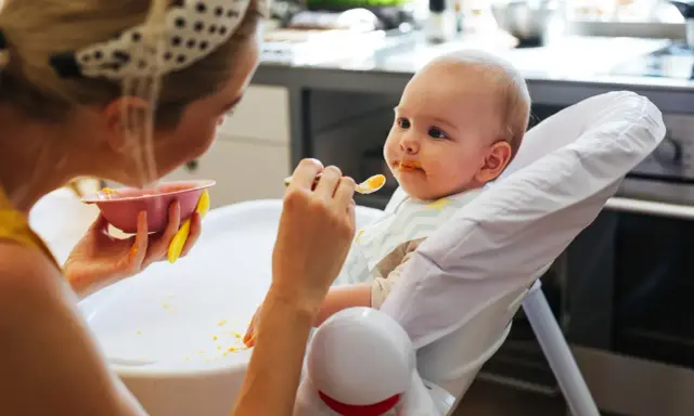 Mama karmi 7 miesięczne dziecko puree z marchewki. Maluch ma pomarańczowe ślady na buzi i śliniaczku.