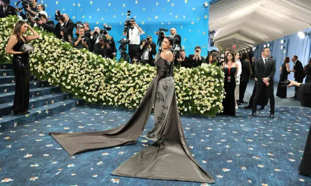 Kylie Jenner arrives at the Met Gala in a dramatic gown, posing for photographers amidst a sea of flowers and a blue, star-studded backdrop.