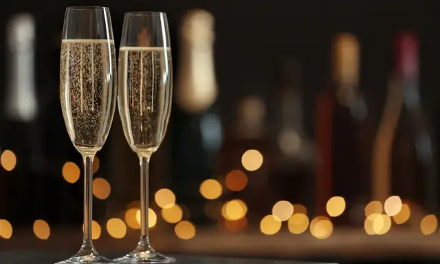 Zwei Sektgläser mit sprudelndem Sekt, im Hintergrund unscharfe Flaschen und Lichter. Ein krug 169 für besondere Anlässe.