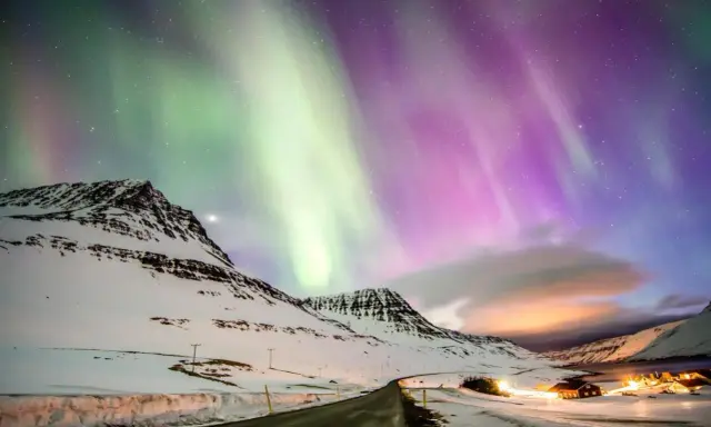 Abisko norrsken målar himlen i grönt och lila över snötäckta berg och en väg som leder mot upplysta hus.