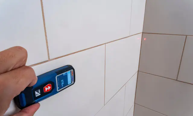 Mano sujetando un aparato para medir con láser rojo apuntando a la esquina de una pared con baldosas blancas.