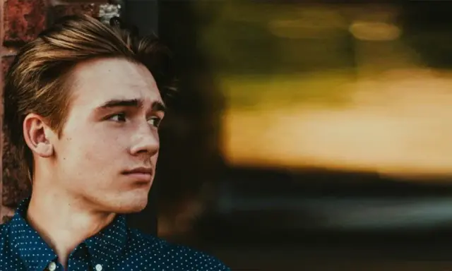 Junger Mann mit stylischen herren frisuren mittellang, lehnt an einer Ziegelwand.