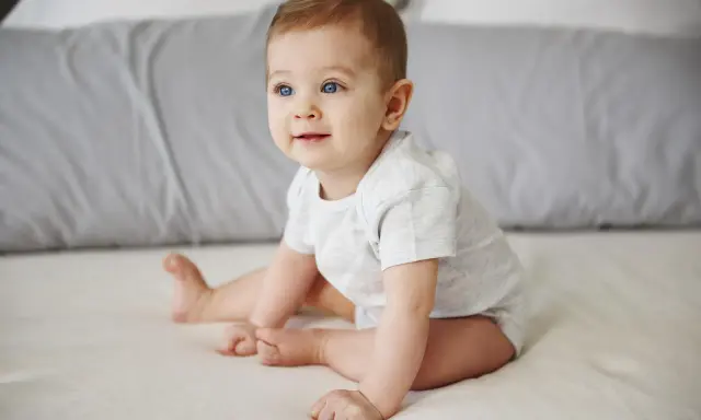Niebieskooki niemowlak w jasnym body siedzi na łóżku, patrząc w bok.