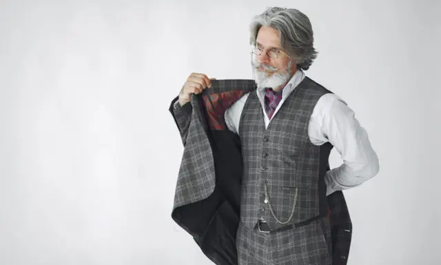 Elegancki mężczyzna z siwą brodą i wąsami, ubrany w garnitur w kratę, prezentuje się jak arbiter elegantiarum.