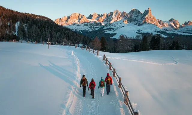 Quattro escursionisti camminano su un sentiero innevato al Passo di Lavazè, con le maestose vette dolomitiche illuminate dal sole al tramonto.