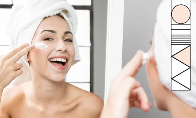 Donna sorridente con asciugamano in testa applica crema sul viso, parte della sua routine skincare.