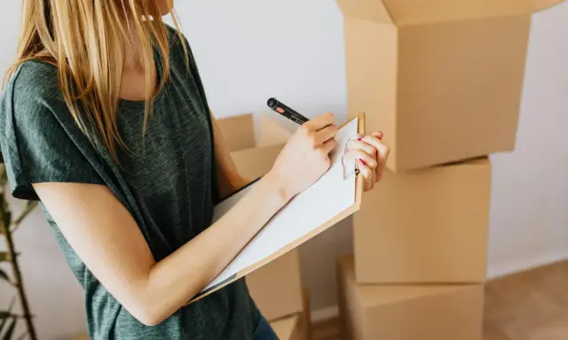Mujer rubia con camiseta verde, revisando una lista en un portapapeles, rodeada de cajas de mudanza. El arte 19 lau inspira la organización.