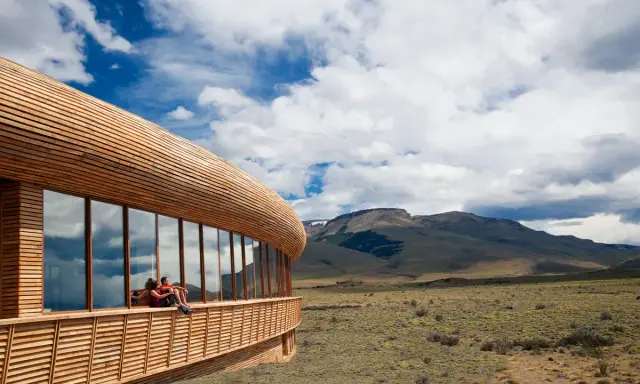 Coppia seduta su una terrazza di **edifici in legno famosi**, con vista su un paesaggio montano e cielo nuvoloso.
