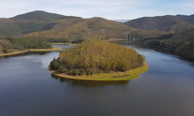 El meandro del Melero, una isla verde rodeada de agua serena, se abre paso entre colinas boscosas bajo un cielo azul.
