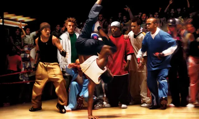 Energetyczna scena z filmów o tańcu. Breakdancer wykonuje akrobację, otoczony przez widzów i innych tancerzy.