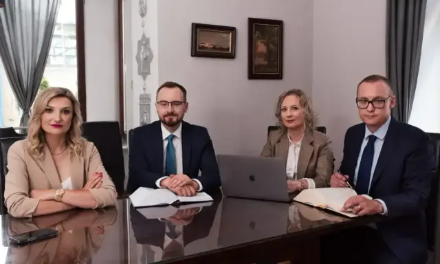 Cztery osoby w strojach biznesowych siedzą przy stole, przed nimi dokumenty i laptop.