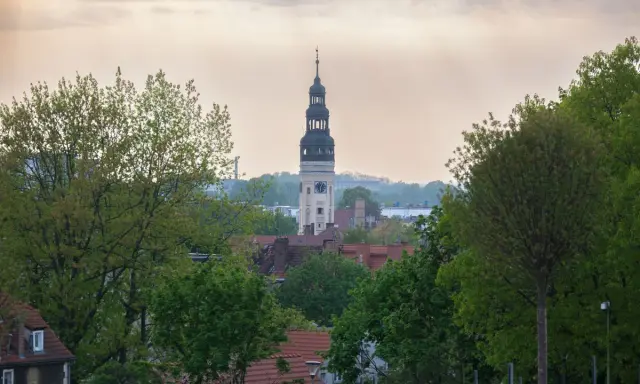 Wieża zegarowa góruje nad zieloną górą, skąd rozciąga się widok na rynek i dachy domów.