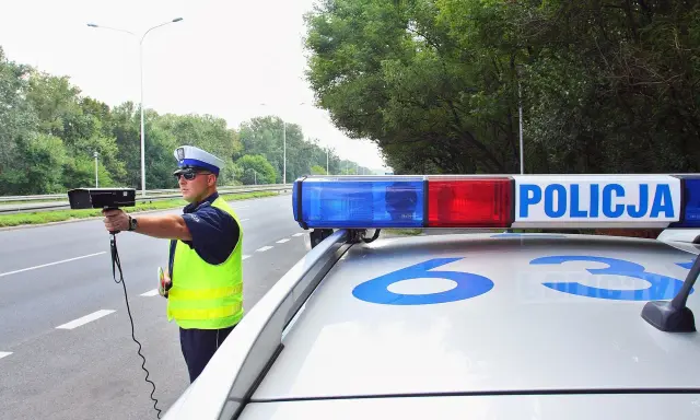 Policjant z radarem na drodze, obok radiowozu z napisem POLICJA.