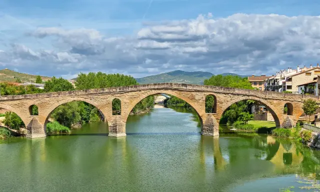 Der historische Puente la Reina überspannt mit seinen steinernen Bögen einen grünen Fluss. Dahinter erstreckt sich eine malerische Stadtkulisse.