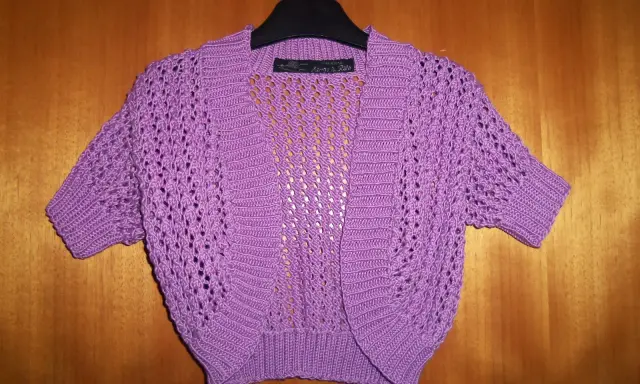 A purple crocheted bolero with short sleeves and an openwork pattern hangs on a hanger against a wooden background.