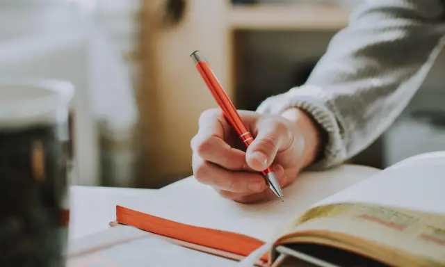 Main tenant une plume rouge, une main écrit dans un carnet. Un synonyme de participer pourrait être collaborer, comme dans cette scène d'étude.