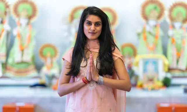 En ung kvinna i traditionella kläder med händerna i bön framför hinduiska statyer, en bild som väcker tankar om mantra hinduism.