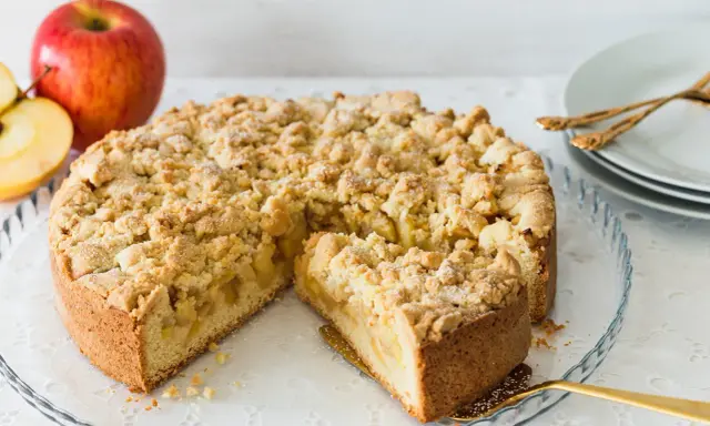 Ein saftiger Apfelkuchen mit Streuseln, ein Stück herausgeschnitten, daneben Äpfel.