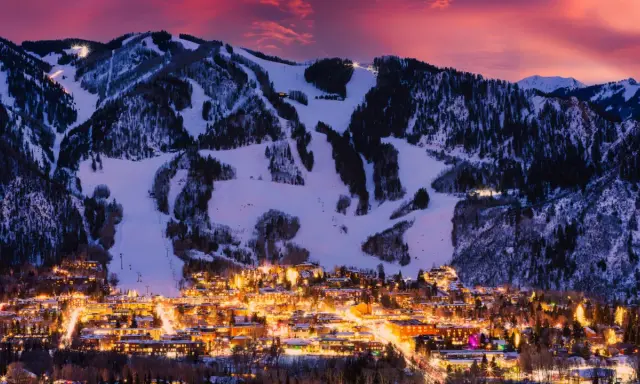 Glitzerndes Aspen, Colorado, im Abendrot. Schneebedeckte Berge mit Skipisten über einer Stadt, die in warmem Licht erstrahlt.
