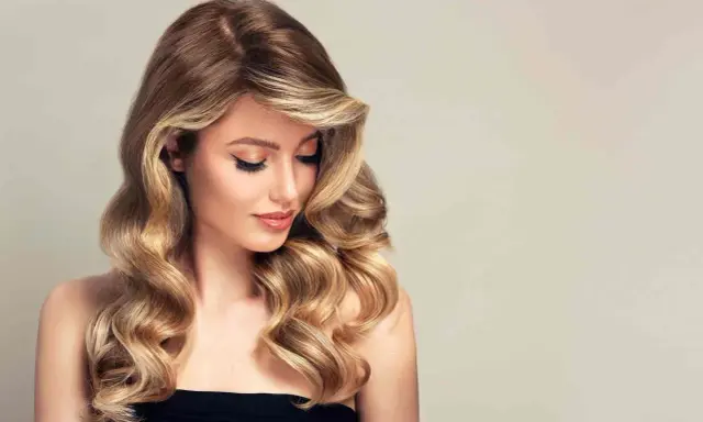Kobieta z falowanymi, brązowo-blond włosami, delikatnym makijażem i spojrzeniem skierowanym w dół.