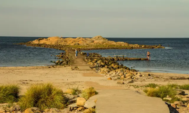 En stenpir leder ut till en klippö. En person går på piren, en annan badar. Perfekt för aktiviteter i Halmstad.
