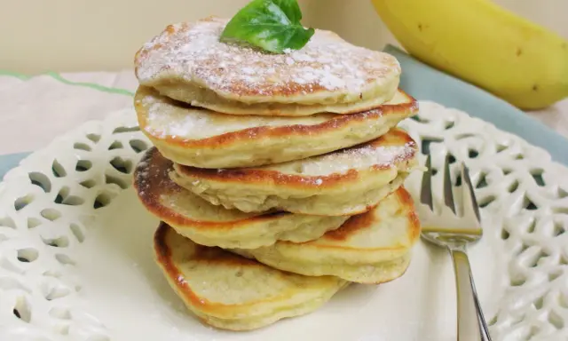 Stos puszystych, trzyskładnikowych placków bananowych, posypanych cukrem pudrem i udekorowanych listkiem, obok dojrzałego banana.