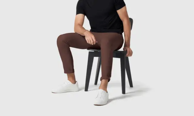 A man in a black shirt and brown pants sits on a black chair, wearing white sneakers.