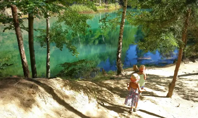 Dwie dziewczynki w słomkowych kapeluszach idą ścieżką nad turkusowym jeziorem w lesie.