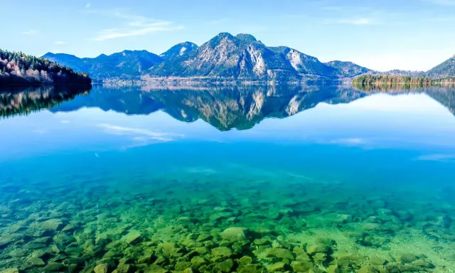 Kristallklarer Badesee in Bayern mit Felsen am Ufer und spiegelnden Bergen im Hintergrund.