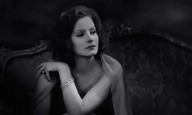 Elegancka Greta Garbo, z błyszczącą bransoletką, spogląda zamyślona z antycznej kanapy.