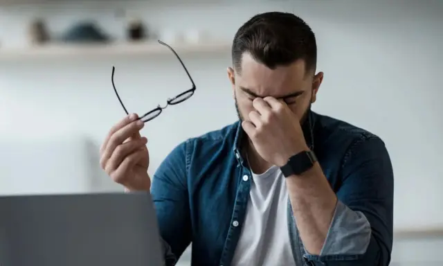 Homme fatigué, lunettes à la main, se frottant les yeux devant un ordinateur. Il est temps de penser à comment protéger ses yeux des écrans.