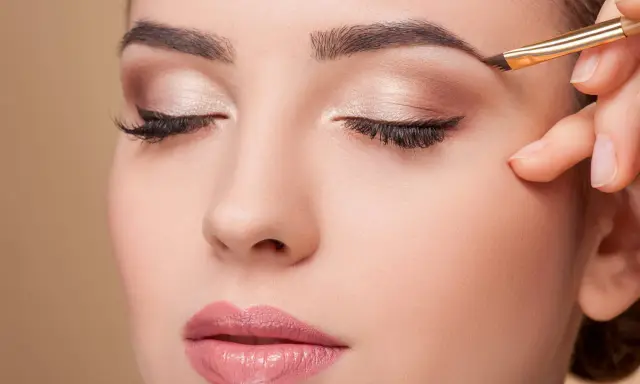 Une main applique un maquillage subtil pour parfaire la forme sourcil d'une femme aux yeux fermés.